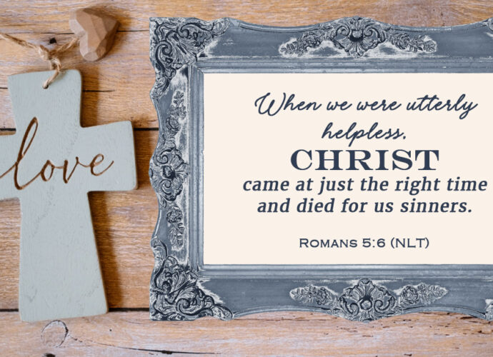 Wooden background featuring a cross with the word "love" engraved and the words of Romans 5:6 within an antique blue frame.