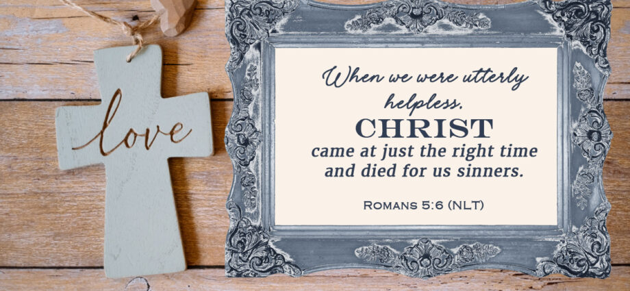 Wooden background featuring a cross with the word "love" engraved and the words of Romans 5:6 within an antique blue frame.